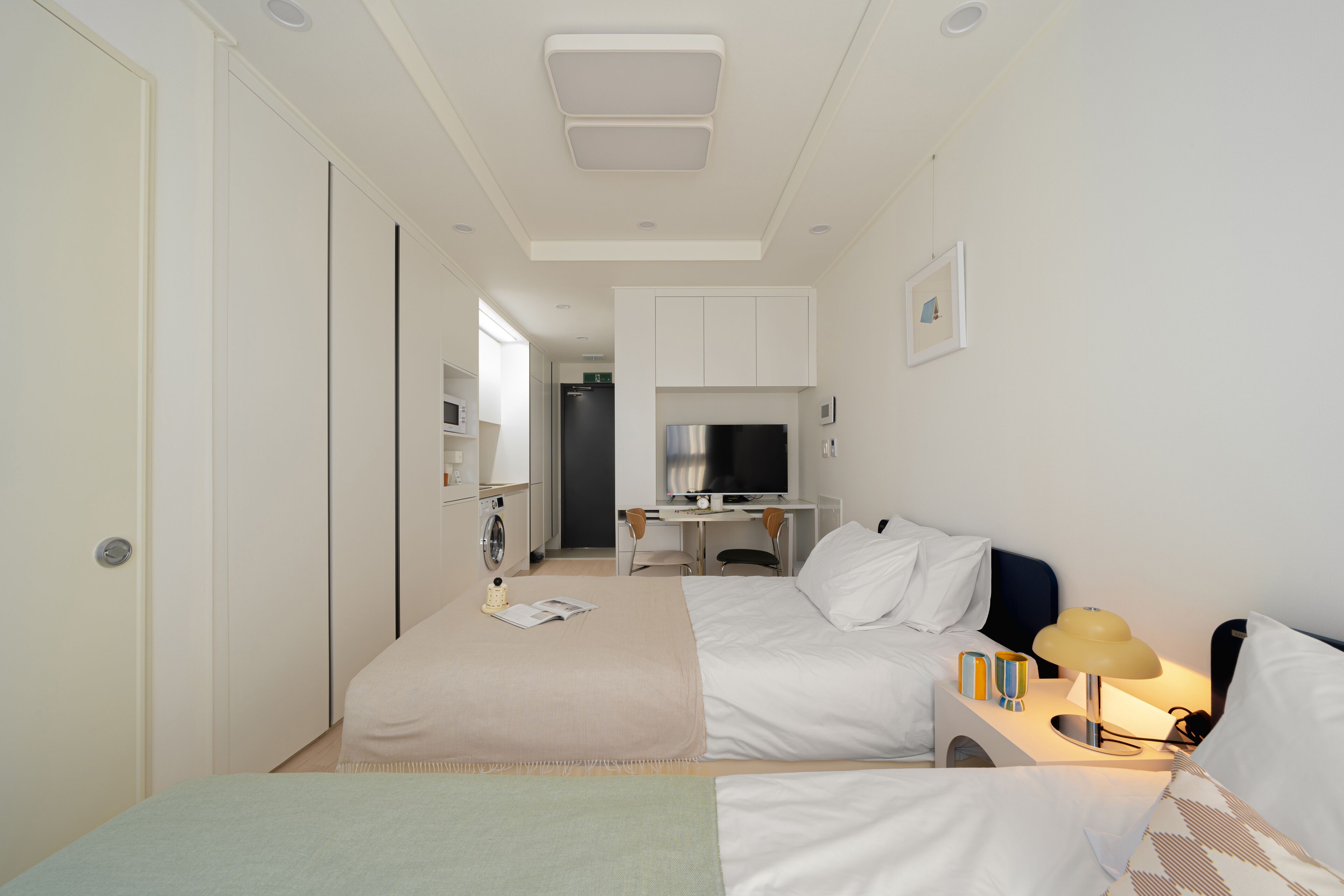 A hotel room with two beds, a desk, a TV, and a kitchenette.