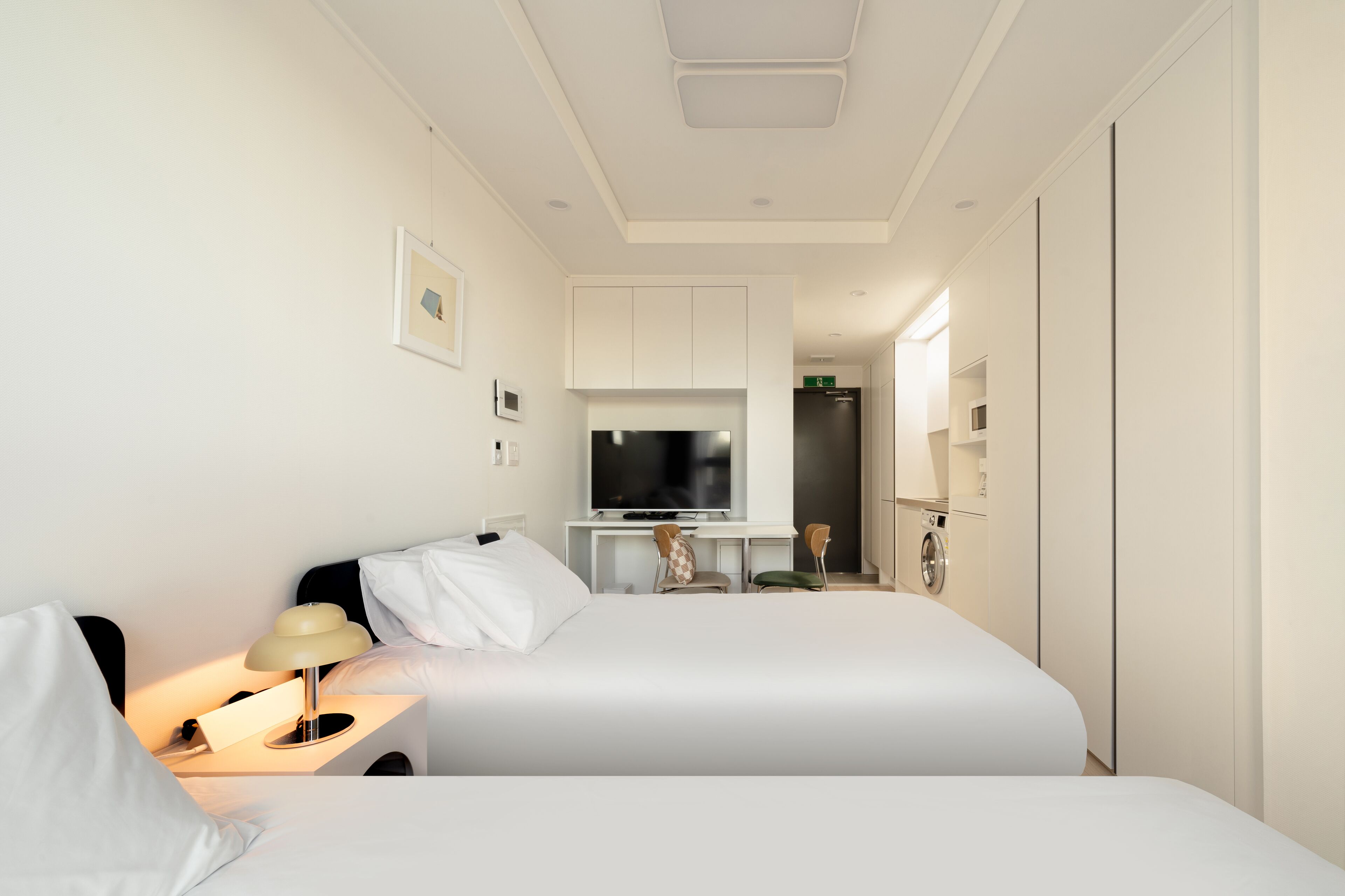 Ein modernes Hotelzimmer mit Bett, Nachttisch, Lampe, Fernseher und einer Küchenzeile.