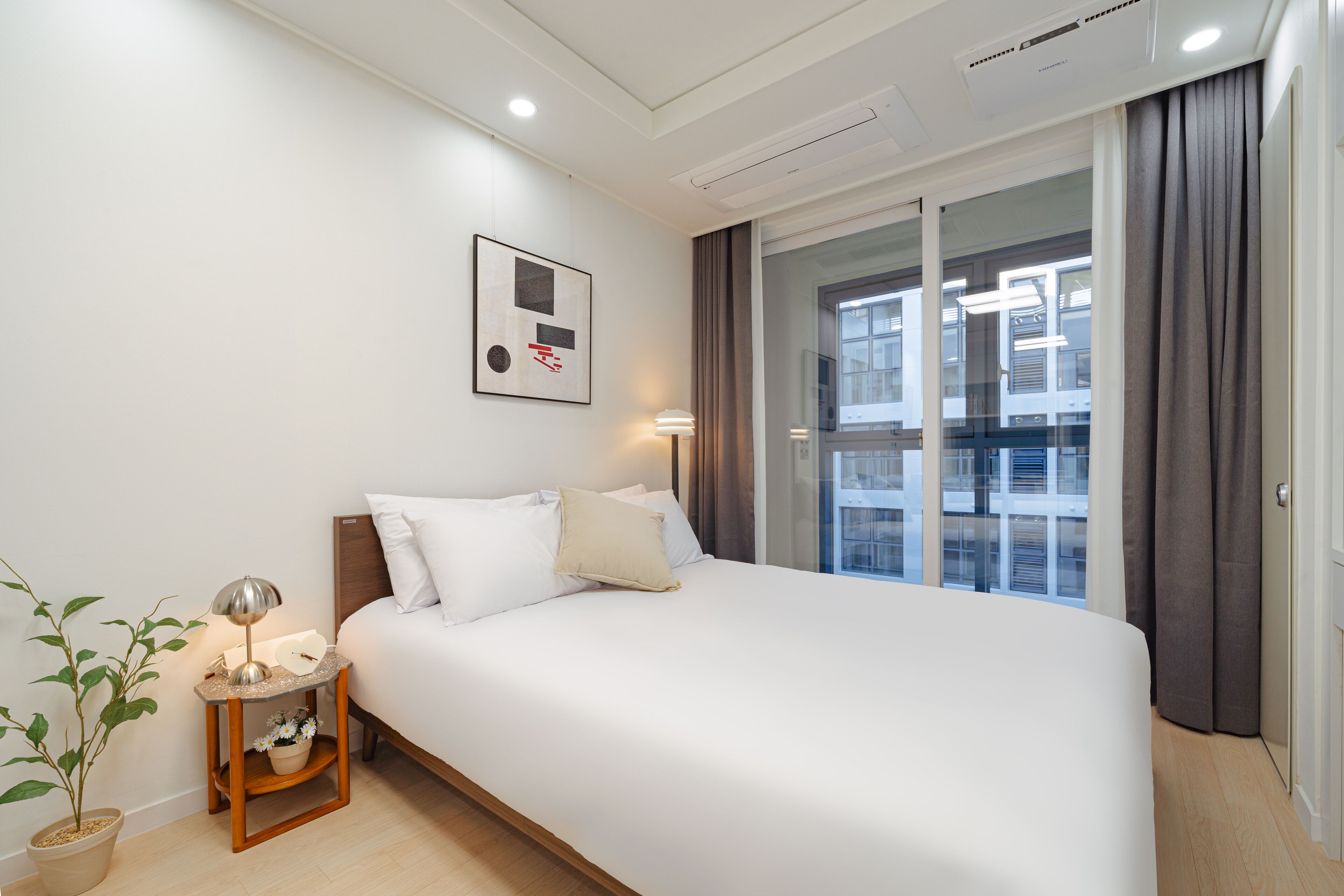 A hotel room with a bed, a bedside table with a lamp, a potted plant, and a window with curtains.