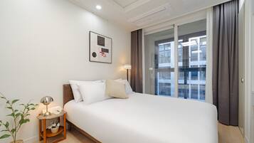 Quarto de hotel com cama, mesa de cabeceira com luminária, planta em vaso e janela com cortinas.