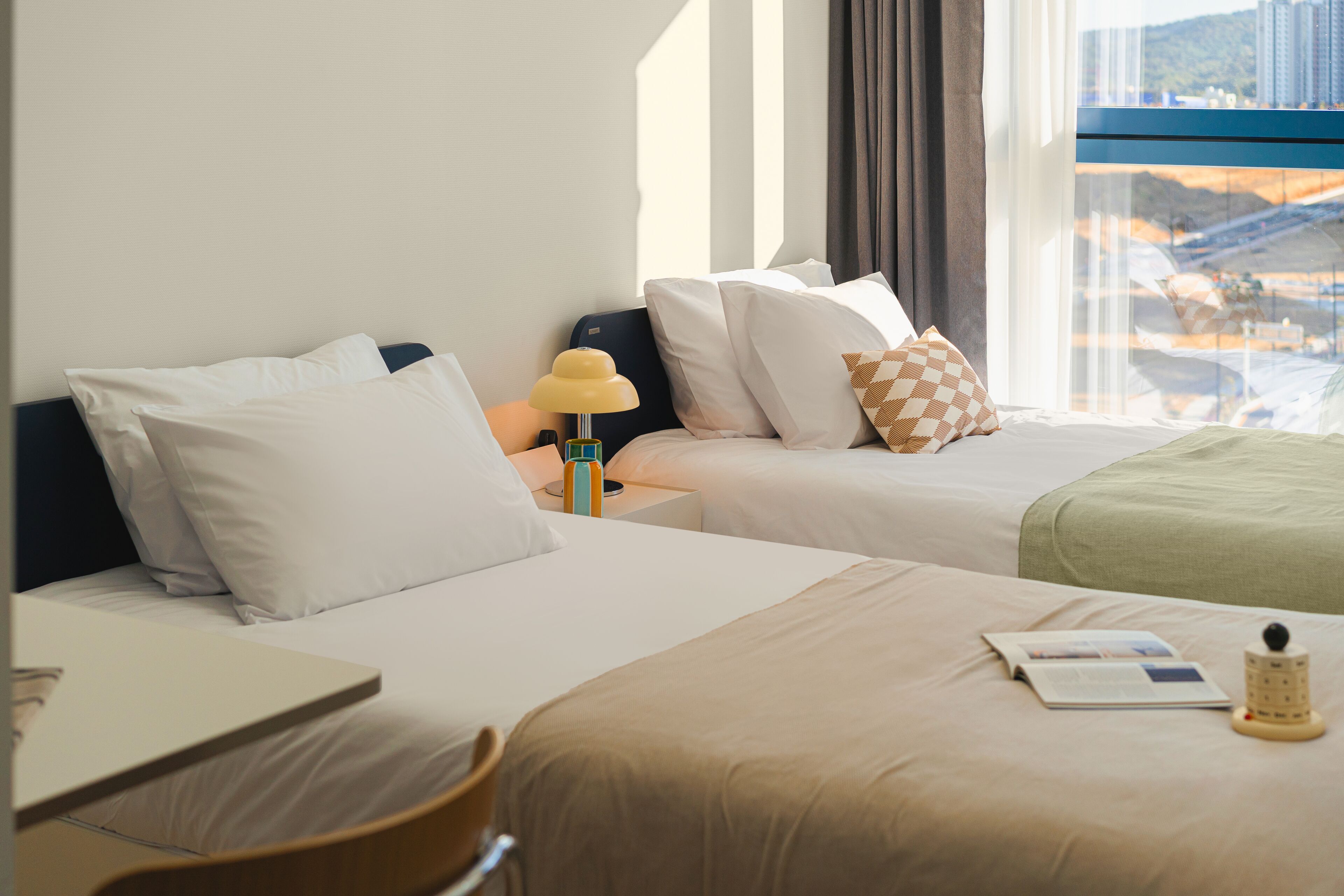 A hotel room with two beds, a desk, and a window with a view of buildings.