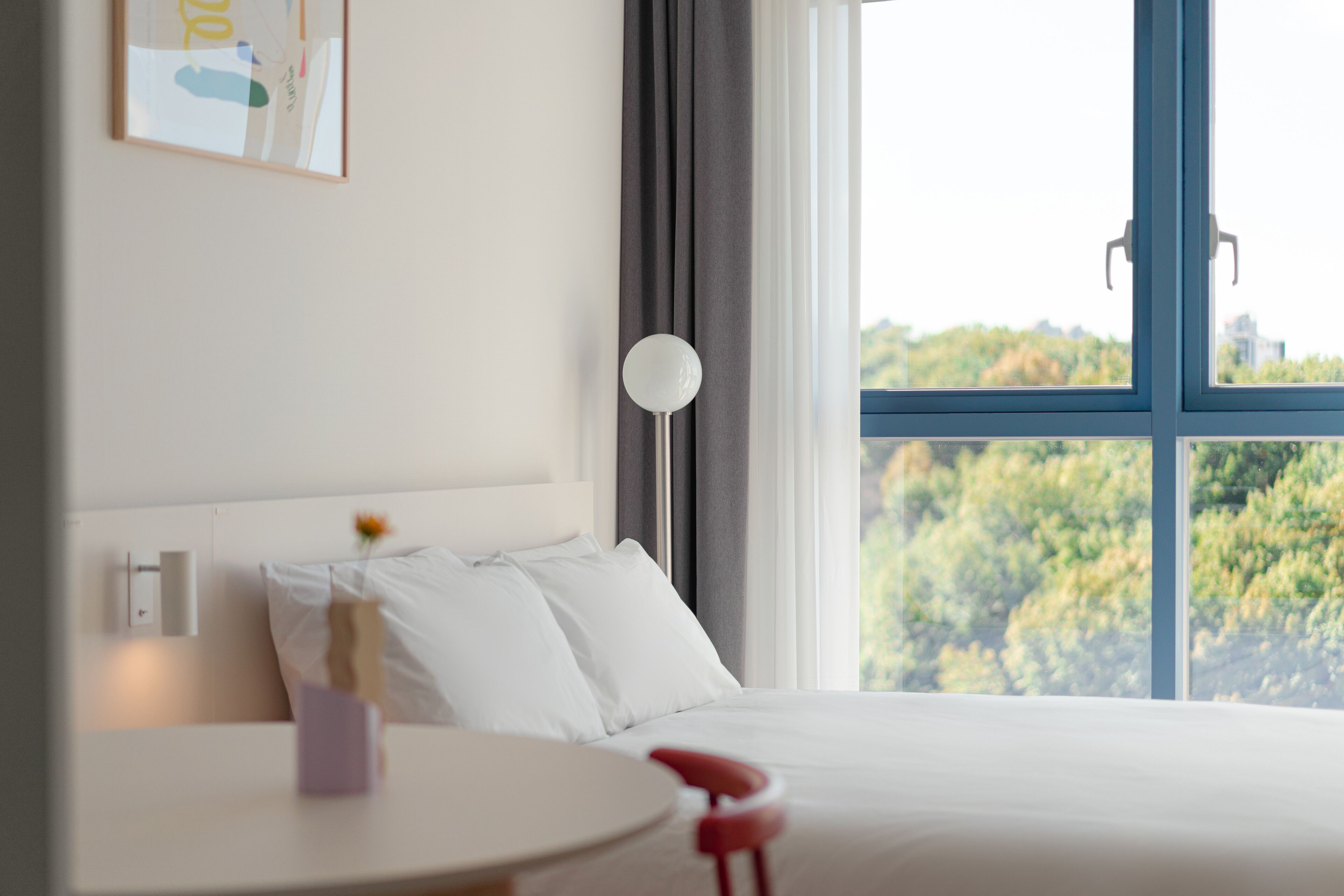 Een hotelkamer met een bed, nachtkastje, lamp en een raam met uitzicht op groen.