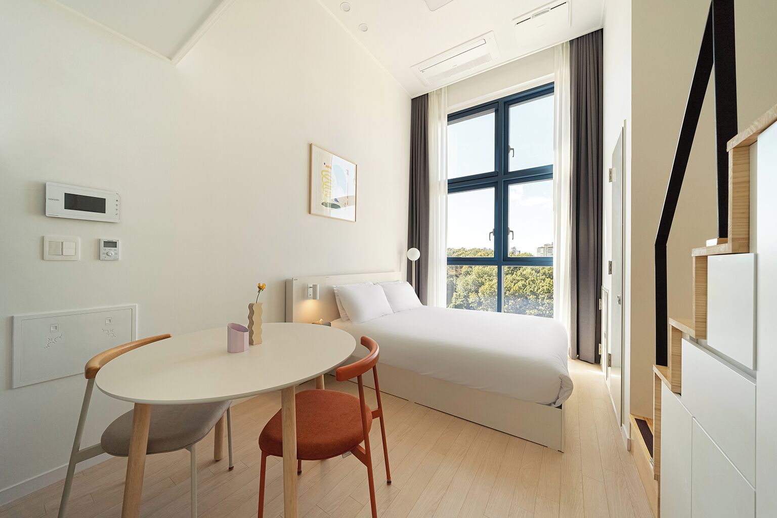 A modern bedroom with a bed, a window with a view, a bedside table, and a chair.