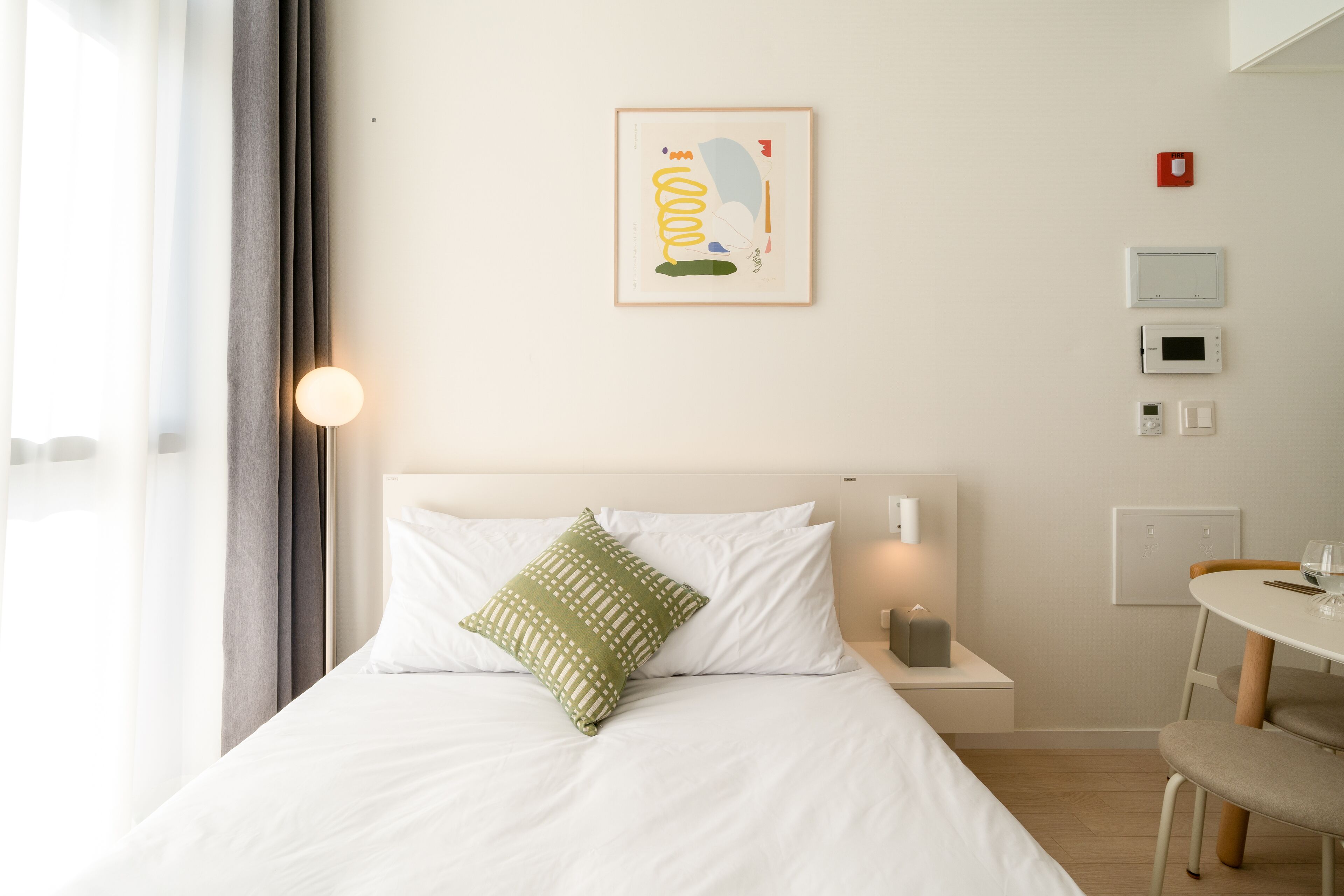 Et hotelværelse med en seng, et natbord med lampe, et lille bord med stol og et vægmonteret termostat.