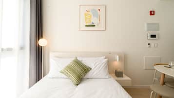 Et hotelværelse med en seng, et natbord med lampe, et lille bord med stol og et vægmonteret termostat.