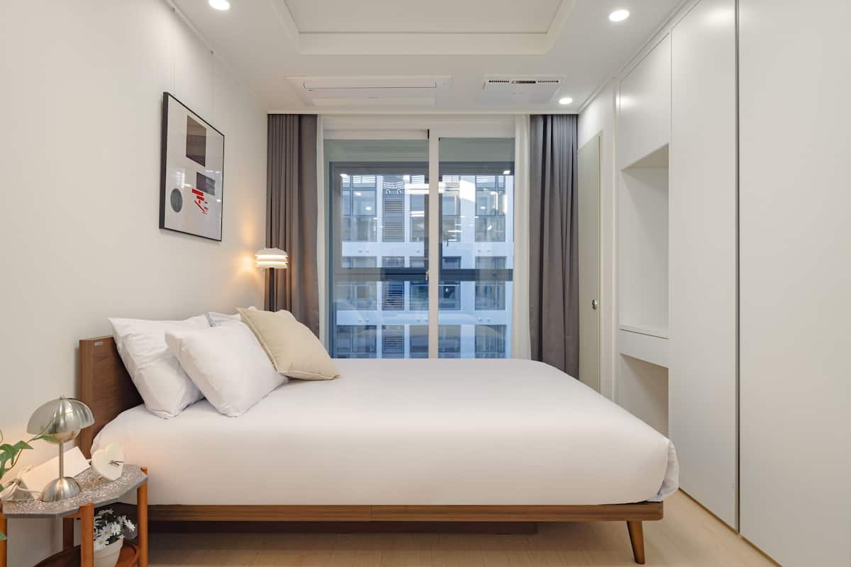 Un dormitorio moderno con una cama grande, una mesita de noche con lámpara, un cuadro enmarcado en la pared y vistas de un edificio a través de la ventana.