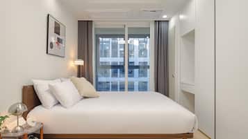 Ein modernes Schlafzimmer mit einem großen Bett, einem Nachttisch mit Lampe, einem gerahmten Bild an der Wand und Blick auf ein Gebäude durch das Fenster.