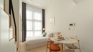 Una habitación de hotel moderna con cama, una mesa de comedor con sillas, una ventana con cortinas y un termostato instalado en la pared.