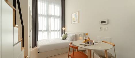 Una habitación de hotel moderna con cama, una mesa de comedor con sillas, una ventana con cortinas y un termostato instalado en la pared.