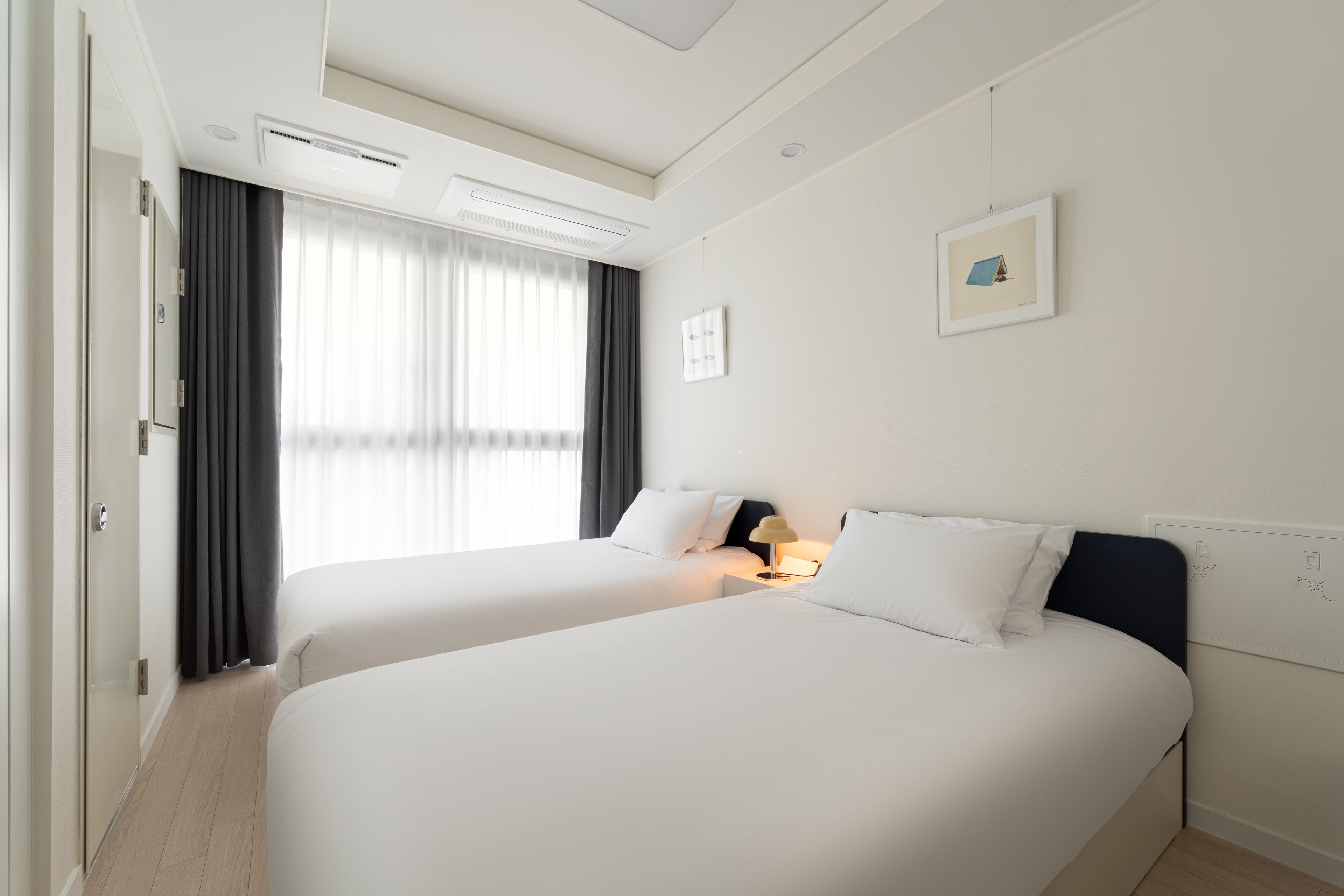 A hotel room with two beds, a window with curtains, a wall-mounted picture, and a ceiling light.