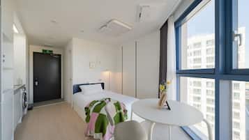 A modern hotel room with a bed, a round table, and a view of the city through large windows.