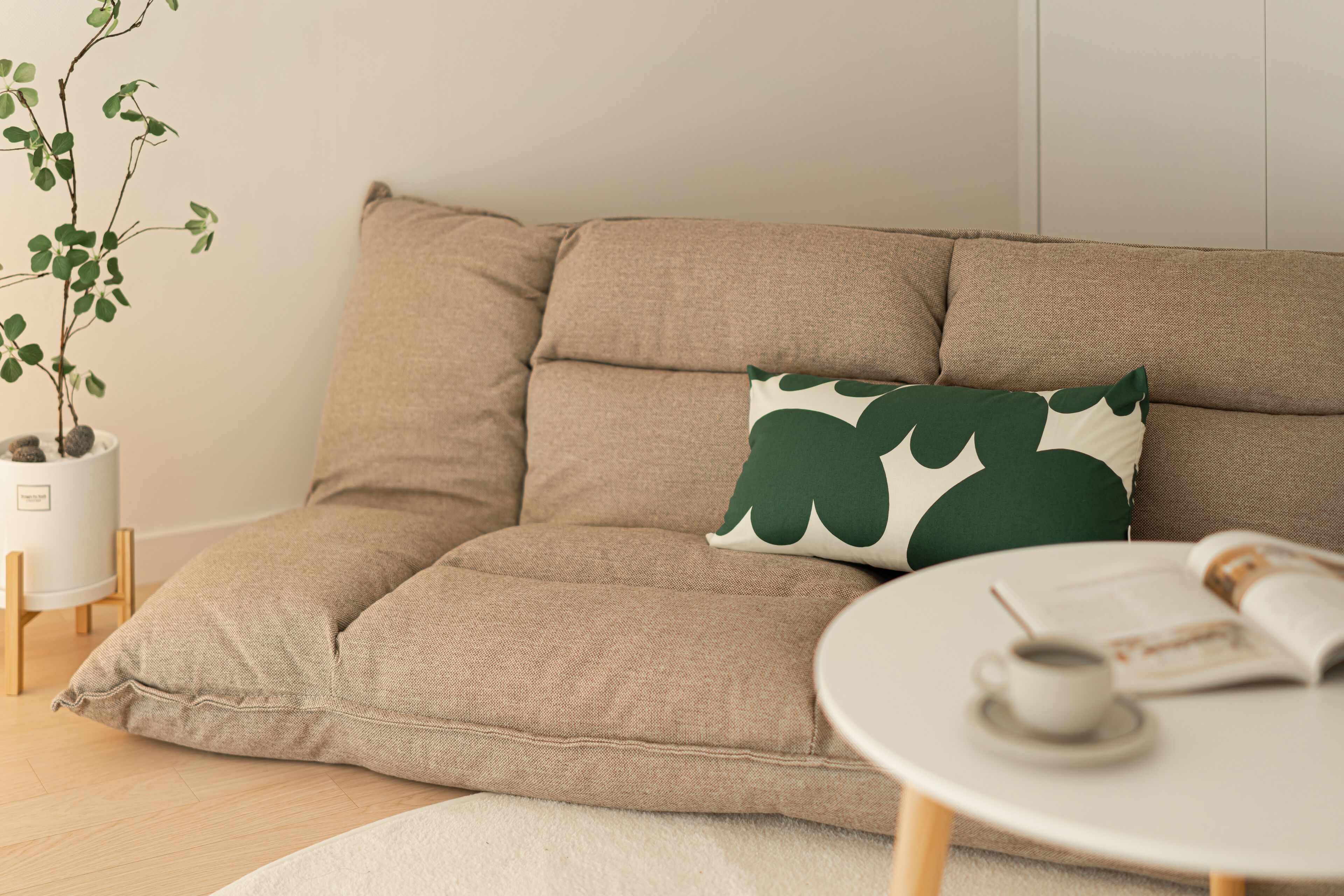 A beige sofa with a green leaf-patterned pillow, a white coffee table with a cup and magazine, and a potted plant.
