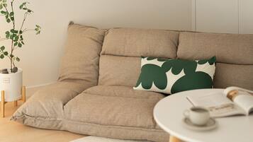 A beige sofa with a green leaf-patterned pillow, a white coffee table with a cup and magazine, and a potted plant.