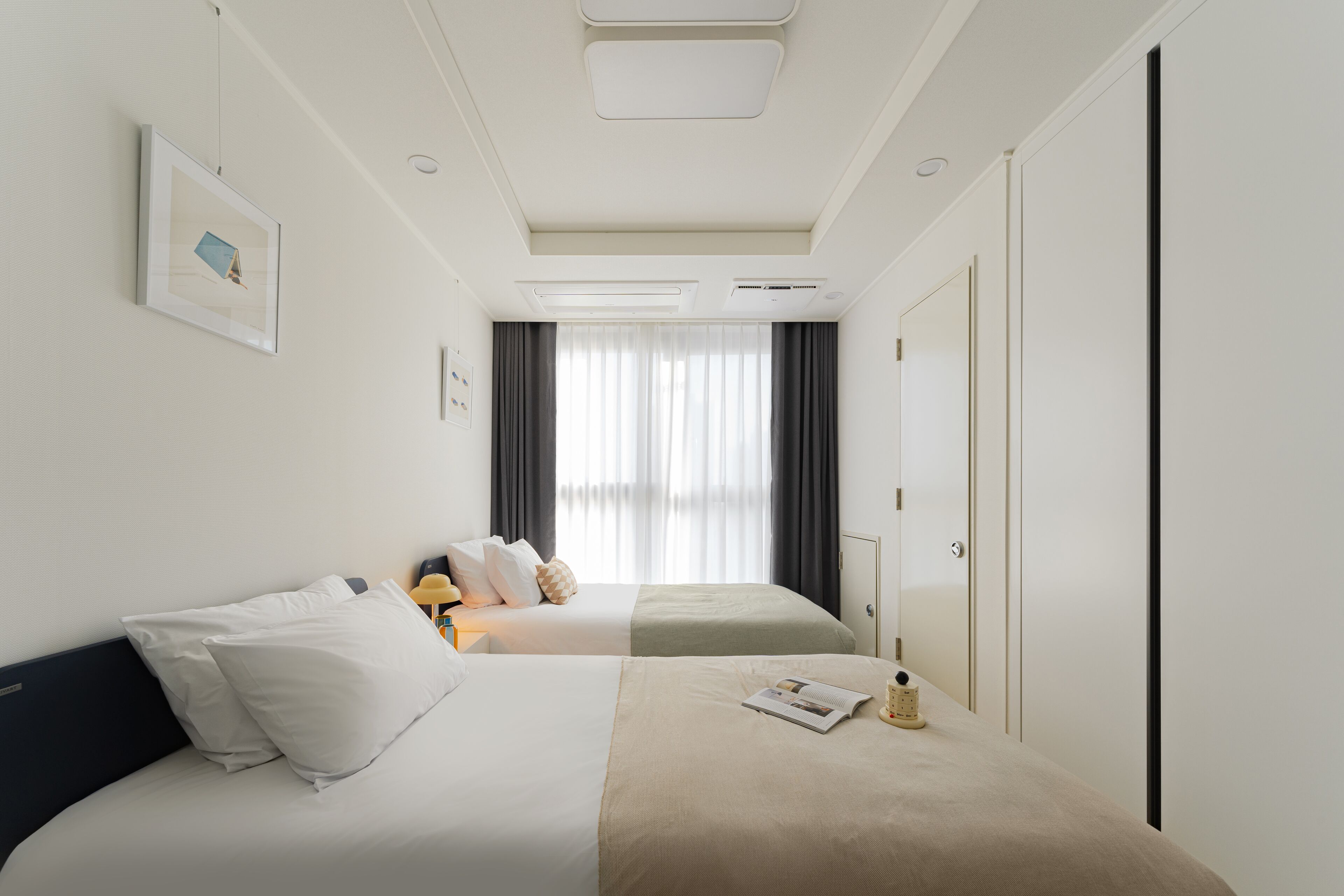 Habitación de hotel con dos camas, un ventanal con cortinas, un cuadro en la pared y una mesita de noche con lámpara y un libro.