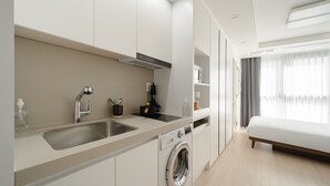 Una habitación de hotel moderna con kitchenette, lavadora, cama y un amplio ventanal con cortinas.