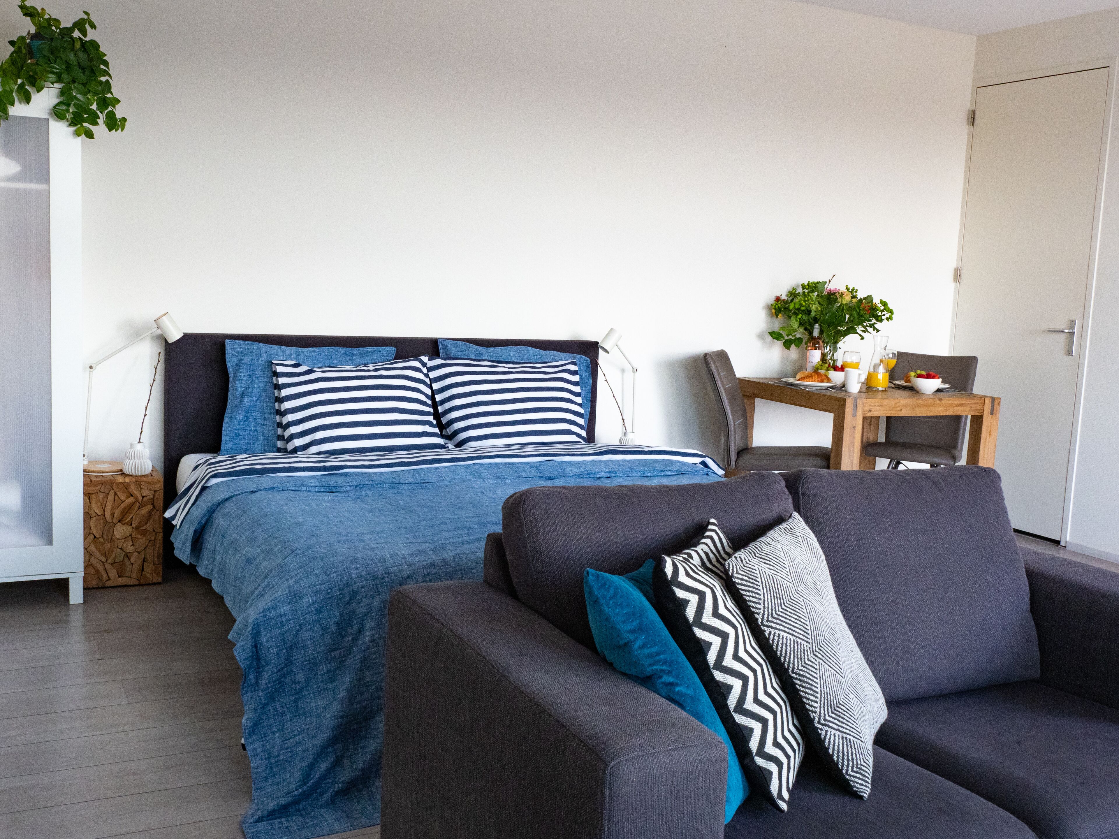 A modern bedroom with a bed, a grey sofa, a wooden nightstand, a table with breakfast, and a door.