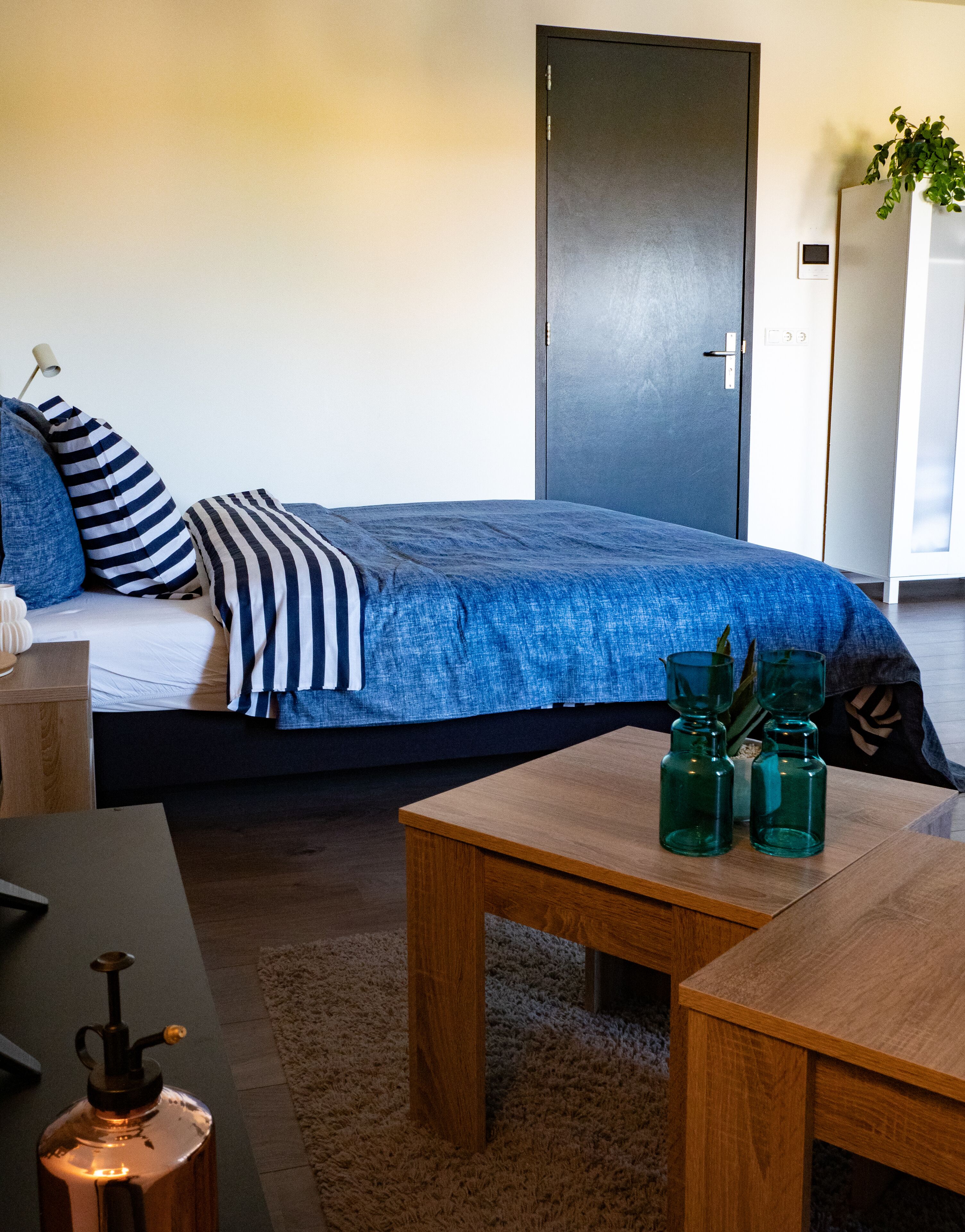 Un dormitorio con cama, mesita de noche y una mesa de madera con botellas de vidrio verde.