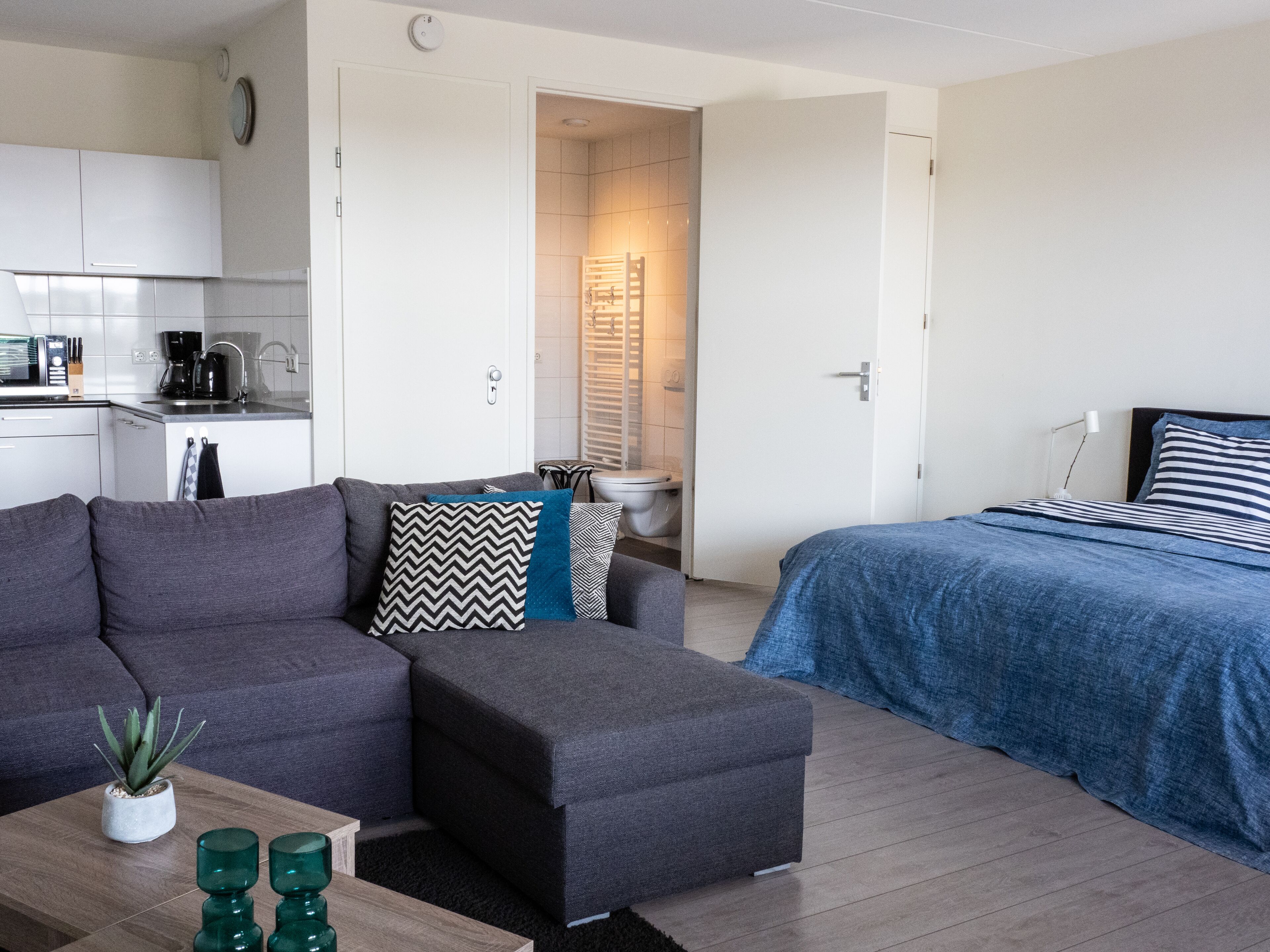 Un espace de vie moderne et compact, comprenant un canapé, un pouf et un lit avec une couvre-lit à rayures. On aperçoit une petite table basse agrémentée d’une plante et de vases en verre vert.