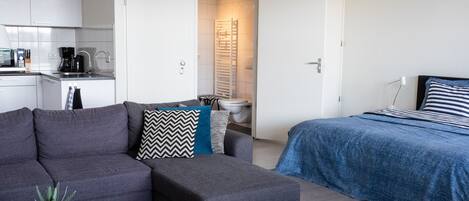 Un espace de vie moderne et compact, comprenant un canapĂ©, un pouf et un lit avec une couvre-lit Ă rayures. On aperçoit une petite table basse agrĂ©mentĂ©e dâune plante et de vases en verre vert.