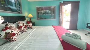 Una cama bien tendida con almohadillas florales, una manta roja y toallas enrolladas. Hay una ventana con vista a una cascada, una lámpara en la mesita de noche y una silla en la esquina.