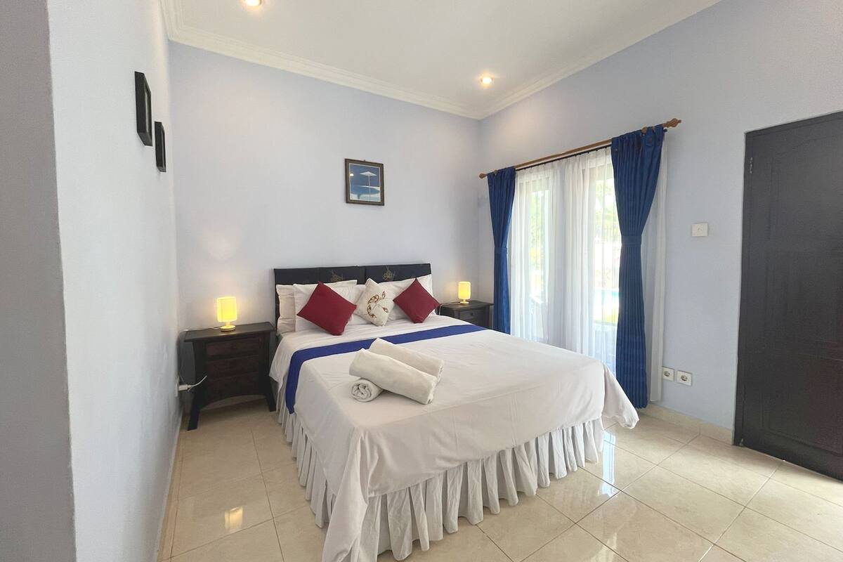 Habitación de hotel con una cama bien hecha, una mesita de noche con lámparas y una ventana con cortinas.