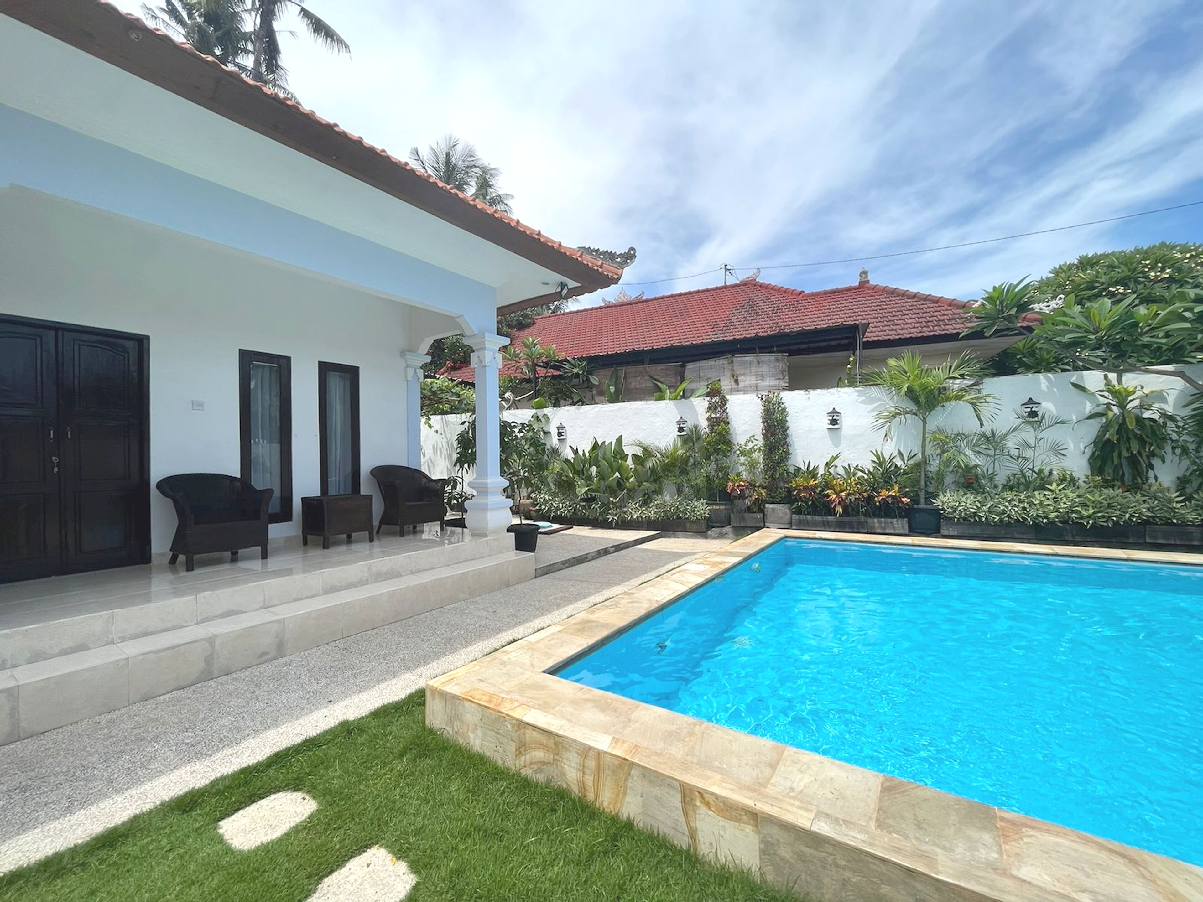 A residential property with a swimming pool, a covered patio with chairs, and a white exterior wall.
