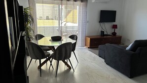 A living room with a glass dining table, black chairs, a grey sofa, a wooden cabinet, a TV, and a plant.