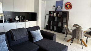 A modern living space with a sofa, a dining table with chairs, a kitchen area, and a bookshelf.