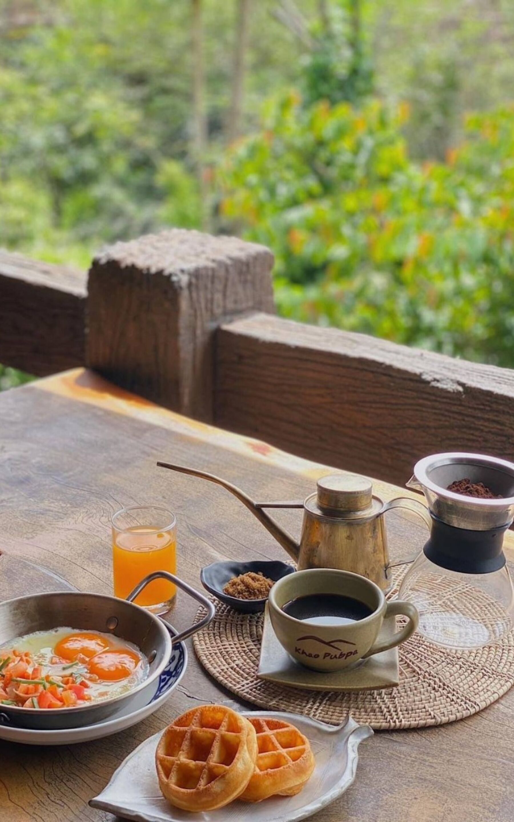 A breakfast setting with waffles, eggs, coffee, and juice on a wooden table with a view of greenery.