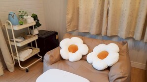 Una habitación con un sofá marrón, dos almohadas grandes con forma de flor, una mesa de centro blanca, una pared blanca, una ventana con cortinas y una estantería blanca con diversos objetos.