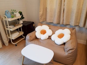 Une pièce avec un canapé marron, deux grands coussins en forme de fleur, une table basse blanche, un mur blanc, une fenêtre avec des rideaux et une étagère blanche sur laquelle sont disposés divers objets.