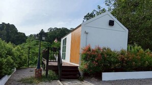 Un pequeño edificio de una sola planta con puerta de madera y fachada blanca, rodeado de vegetación y un camino de grava.
