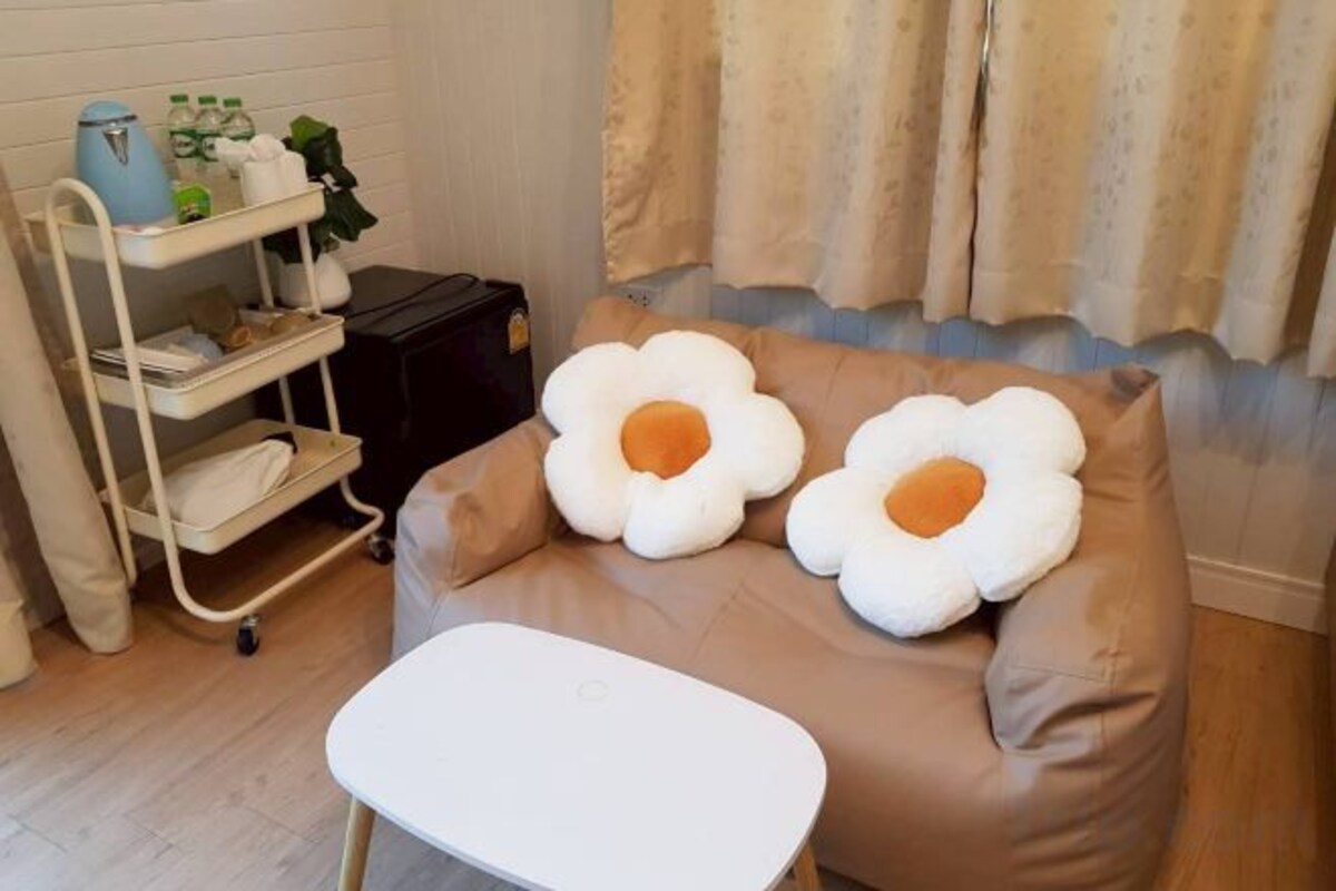 A room with a brown sofa, two large flower-shaped pillows, a white table, a metal cart with items, and a window with curtains.