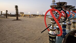 A hookah with a large silver bowl and multiple hoses, set on a sandy surface with palm trees and a building in the background. - Easy Trip Desert Camp (Al Awir)