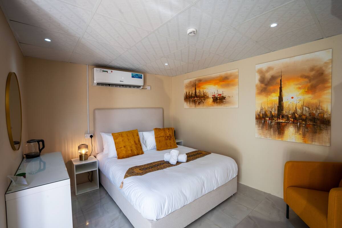Ein Hotelzimmer mit Bett, Nachttisch, Lampe, Klimaanlage und Kunstwerken an den Wänden.