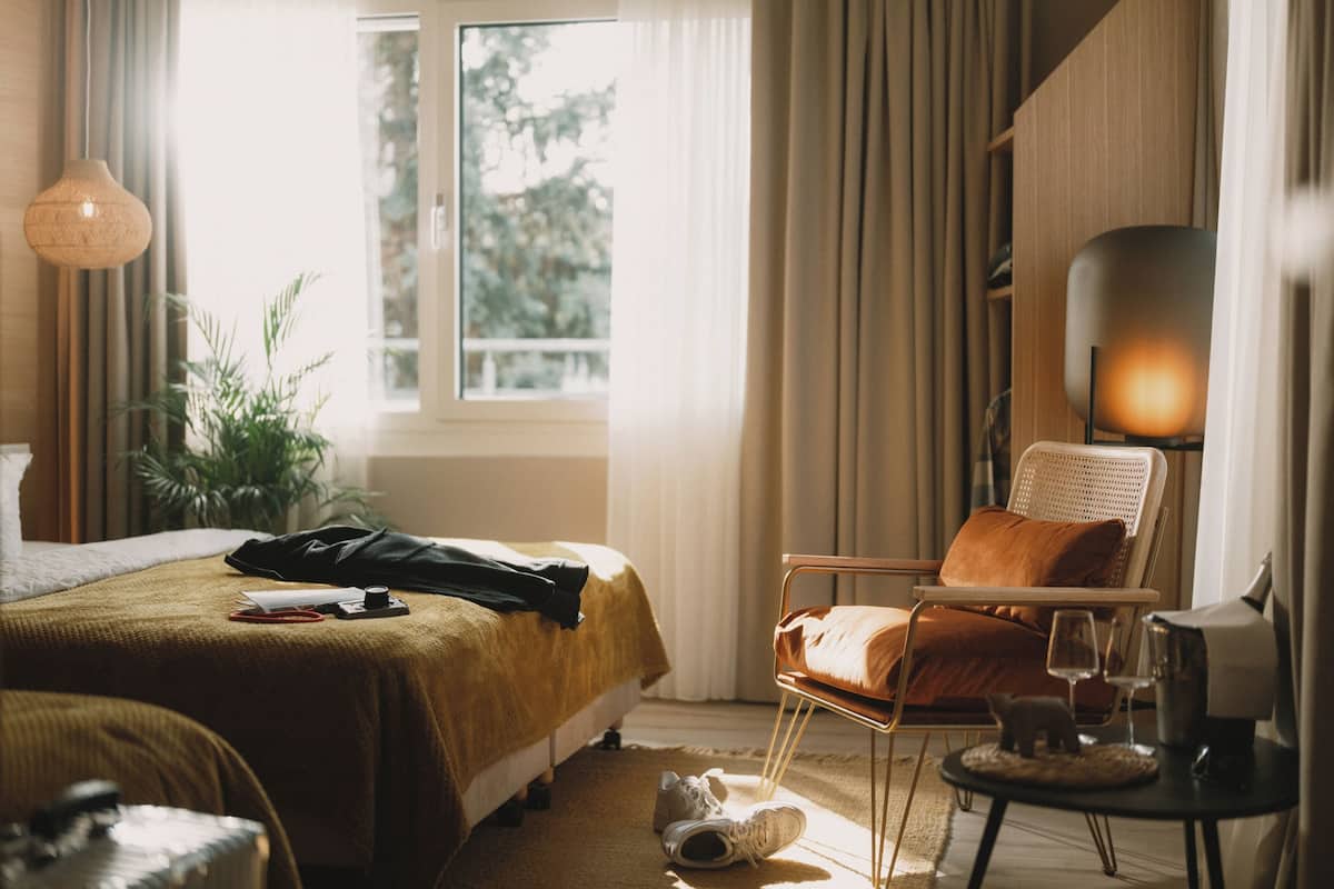 Una habitación de hotel con cama, una silla, una mesa con lámpara y ventana con cortinas.