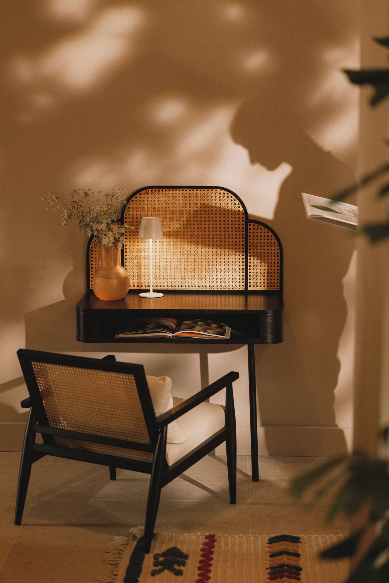 Une pièce comprenant un fauteuil en rotin, une petite table avec un vase et une lampe, et une étagère murale sur laquelle est posé un livre.