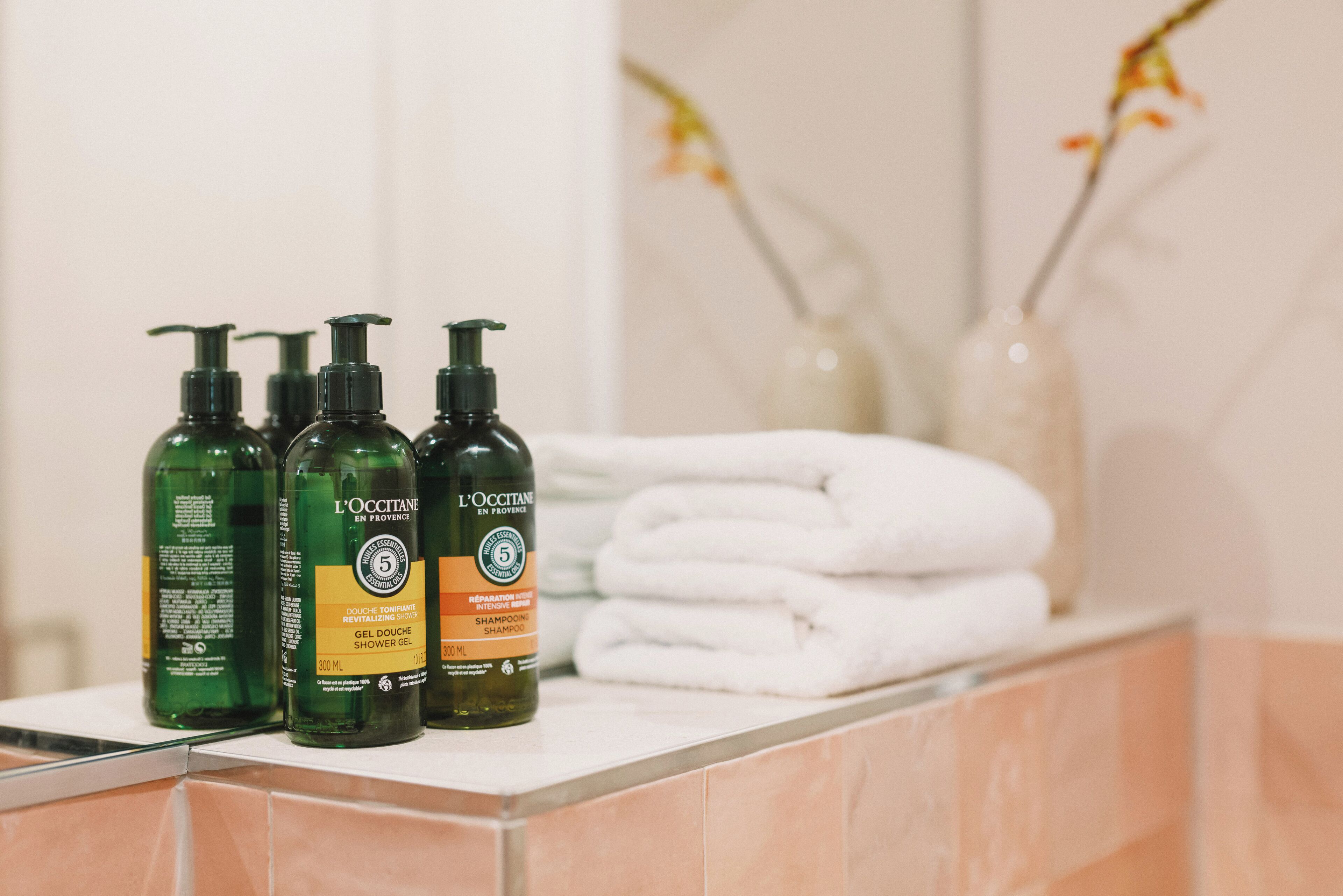 Una encimera de baño con tres botellas de gel de ducha y champú L'OCCITANE, y una pila de toallas blancas.