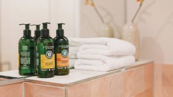 Una encimera de baño con tres botellas de gel de ducha y champú L'OCCITANE, y una pila de toallas blancas.
