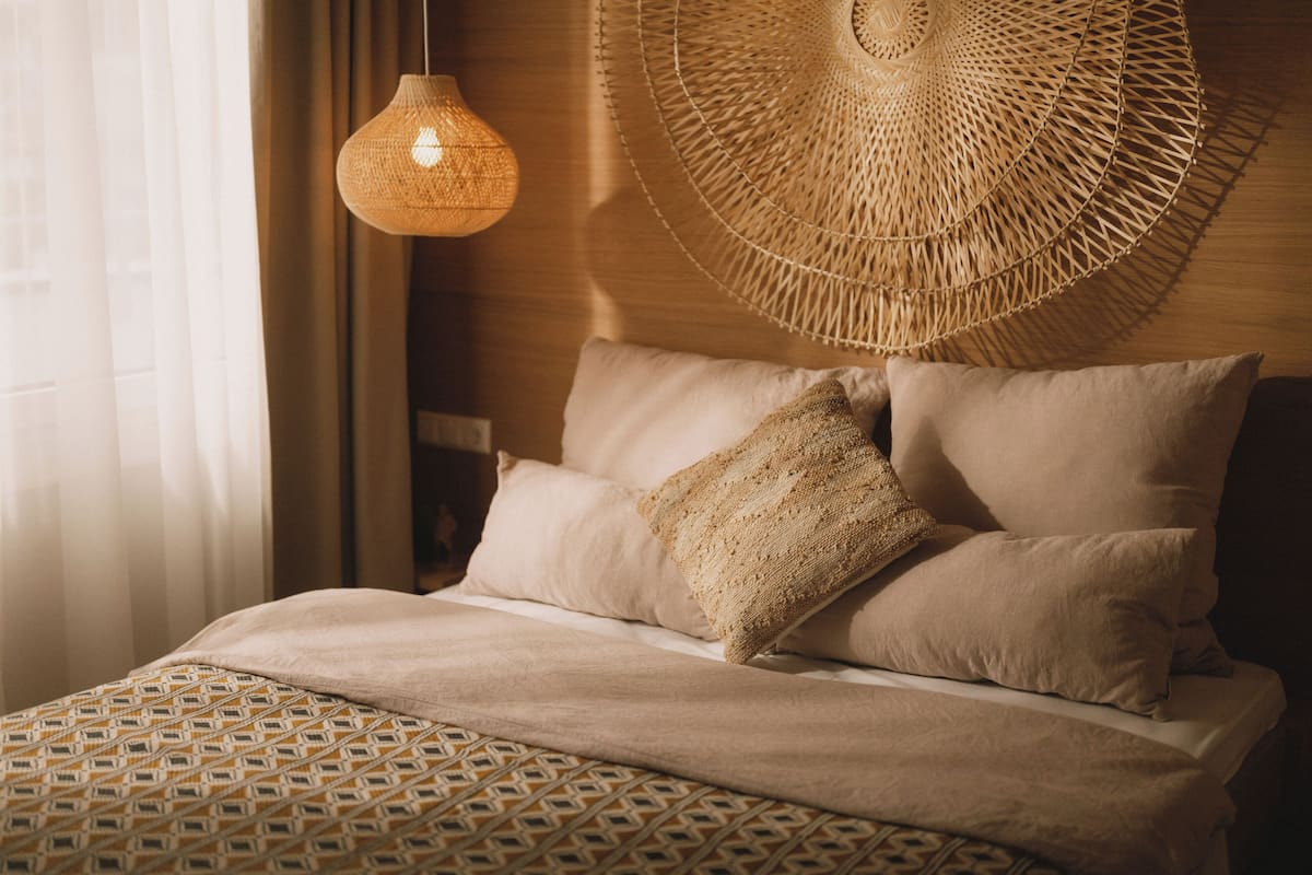 Una cama bien hecha con ropa de cama estampada, un tapiz de pared tejido y una lámpara colgante.