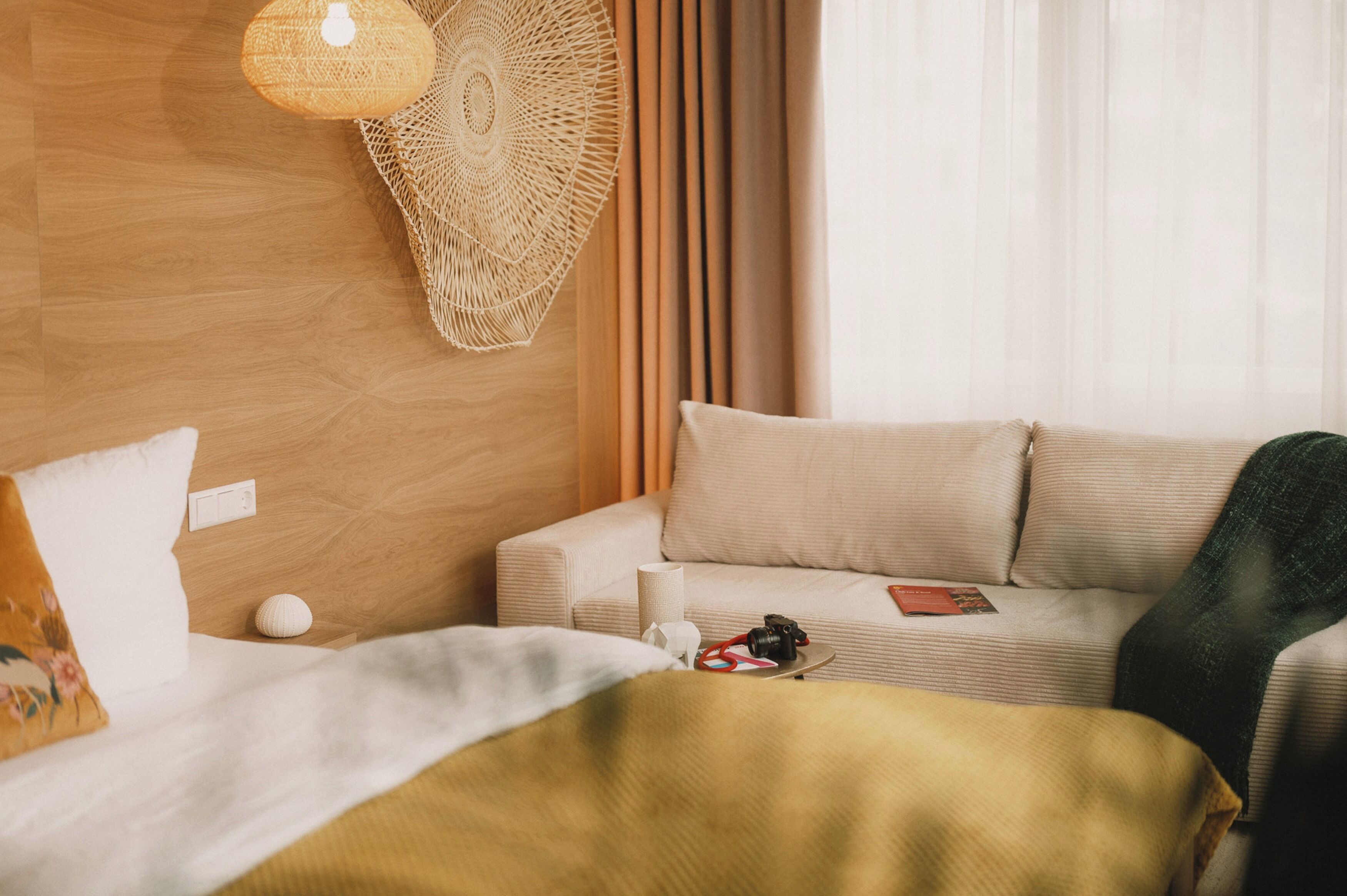 Una habitación de hotel con cabecera de madera, un sofá color beige, una cama con manta amarilla, una lámpara de pared y una ventana con cortinas translúcidas.