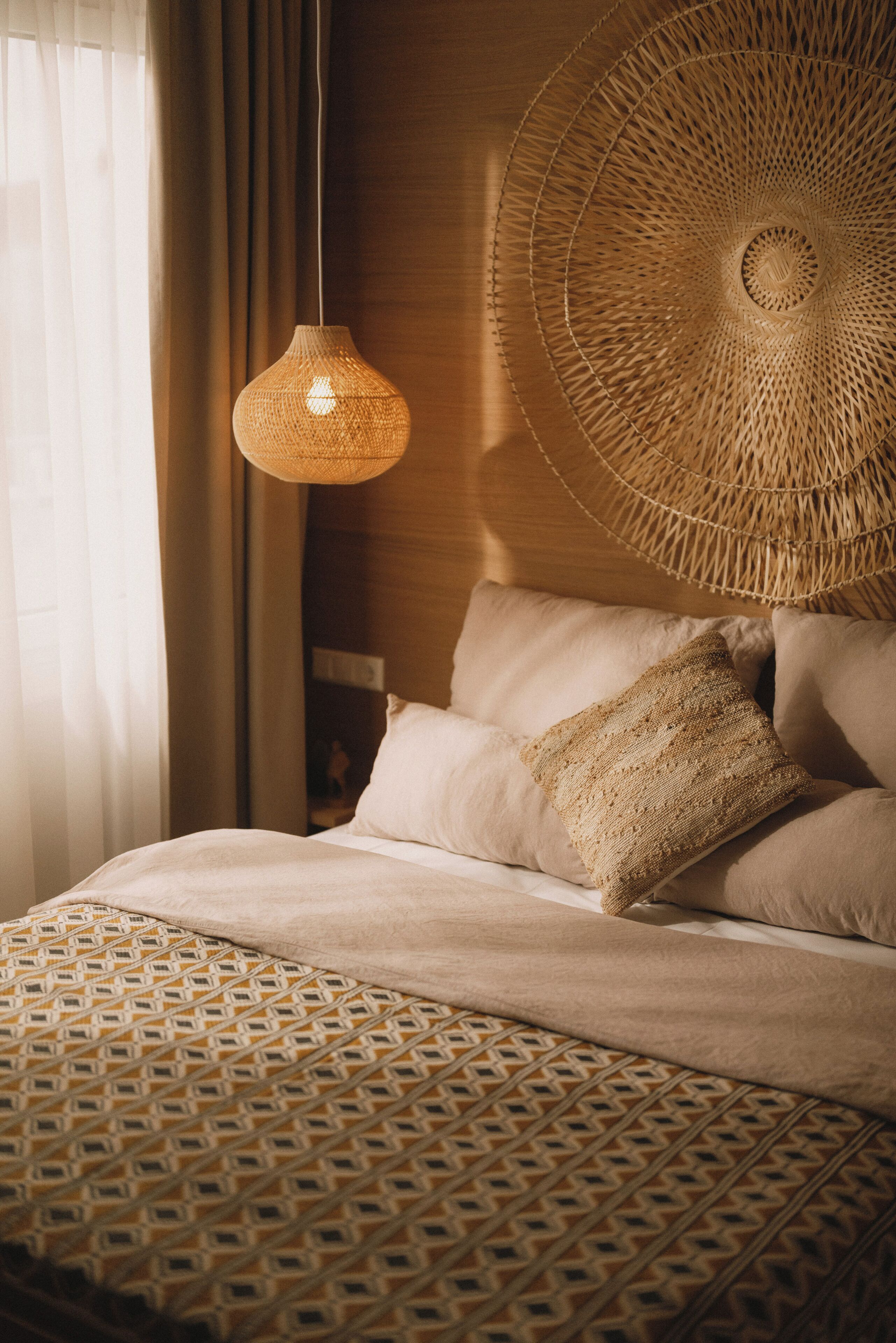 Una cama con una colcha estampada, una lámpara colgante tejida y un cojín con textura.