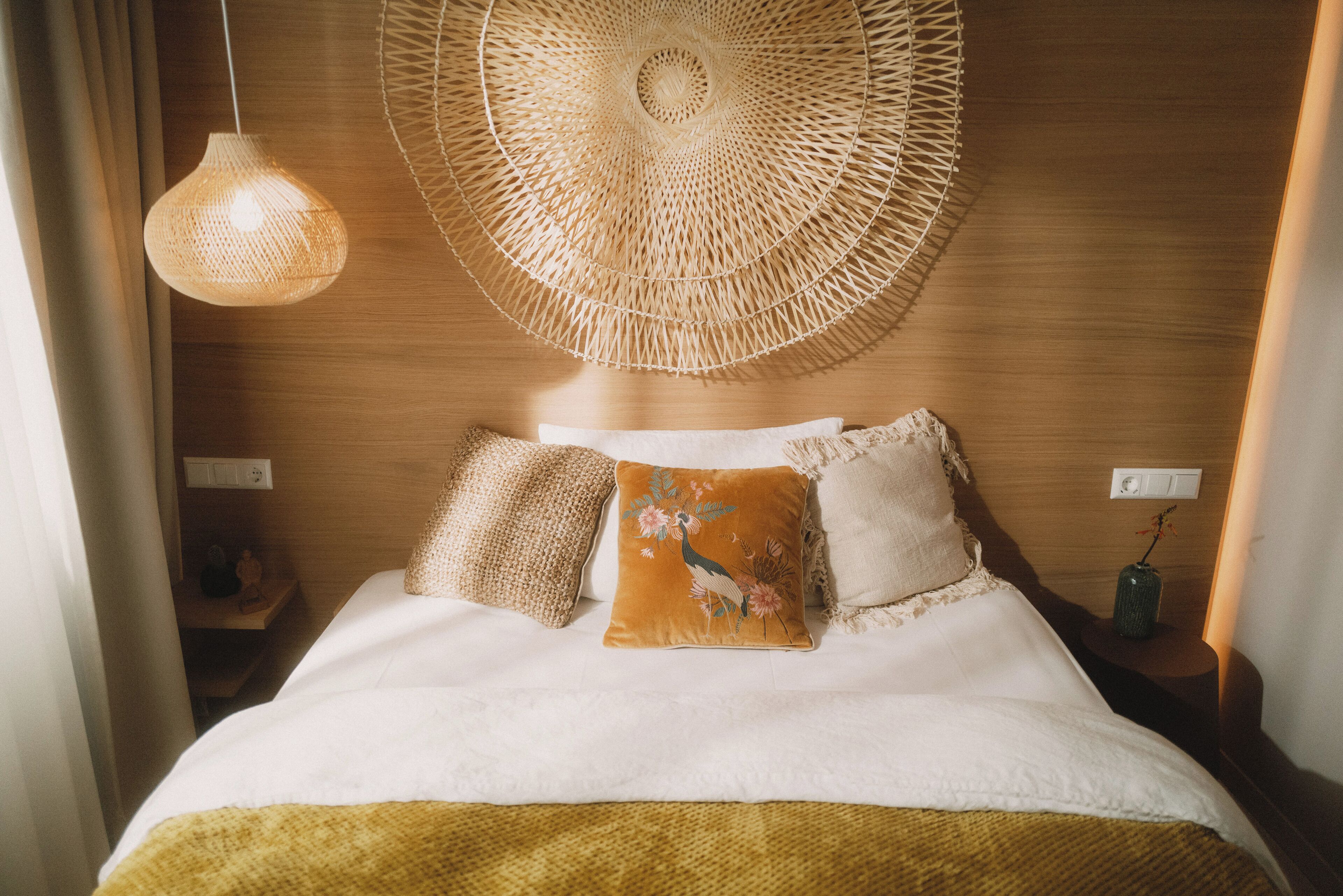 A neatly made bed with a white comforter and two decorative pillows, a woven wall hanging above, and a hanging light fixture.