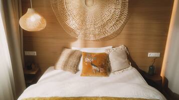 A neatly made bed with a white comforter and two decorative pillows, a woven wall hanging above, and a hanging light fixture.
