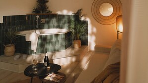 A modern spa room with a bathtub, a small table with wine glasses and a bottle, and a cozy seating area with a large mirror and wall-mounted light fixture. - 2Places Soul (Loerrach)