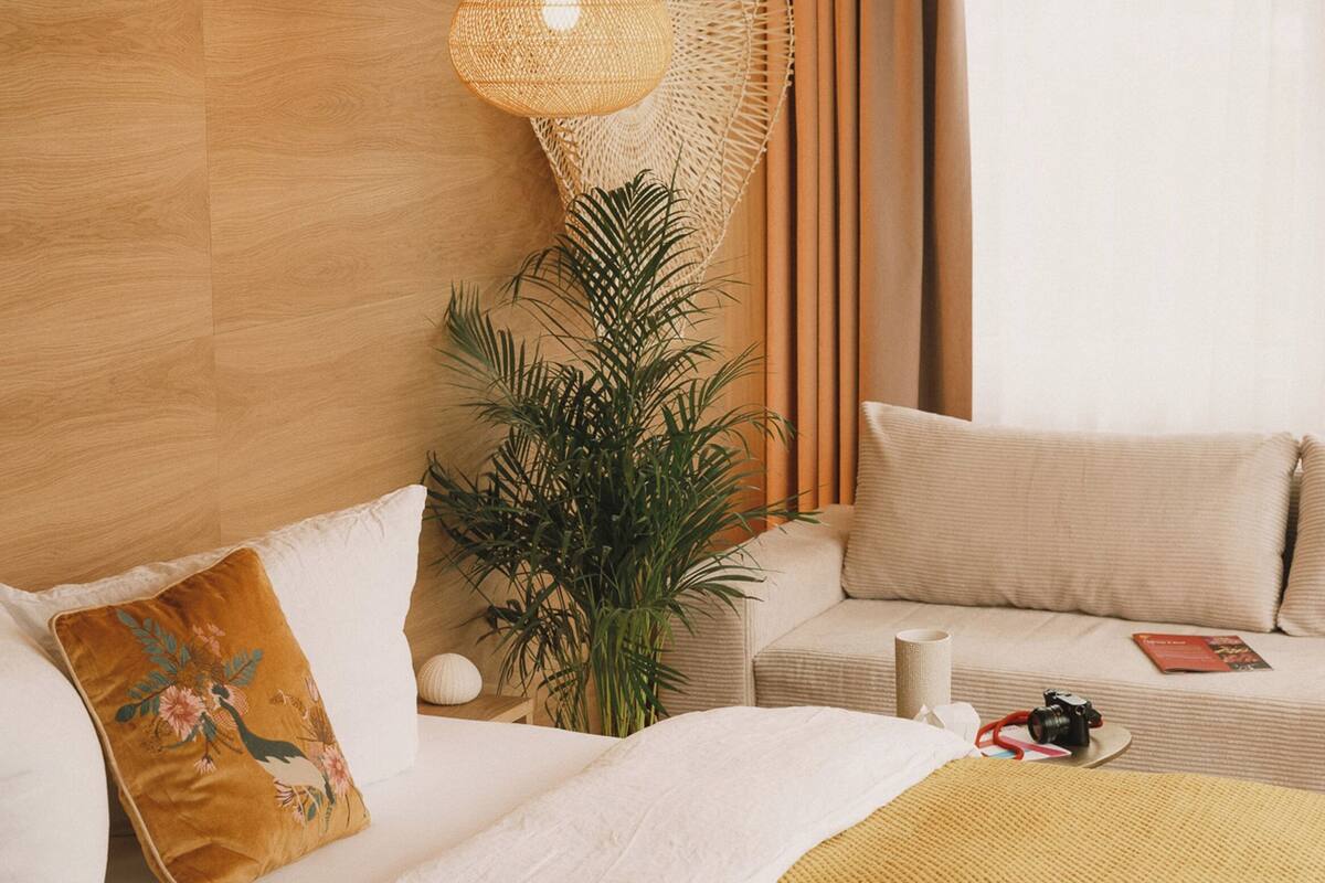 Una habitación de hotel moderna con cabecera de madera, lámpara de techo tejida, cama grande con manta amarilla, almohada blanca y una mesita con una cámara y un libro.