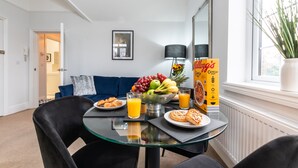 Un coin repas avec une table en verre, des chaises noires, un saladier de fruits, des assiettes de nourriture et une boîte de céréales.