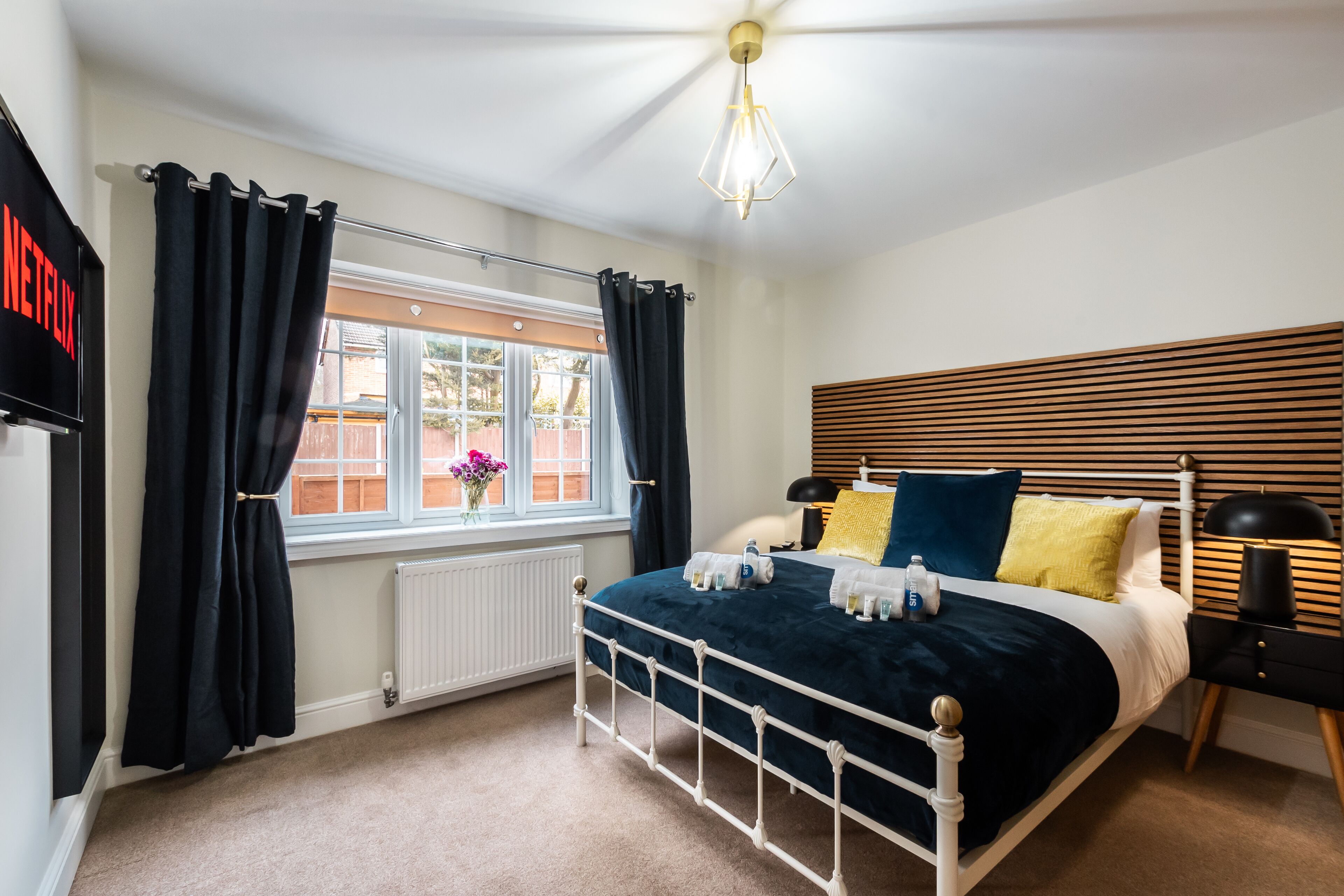 Un dormitorio con estructura de cama metálica blanca, colcha azul oscuro y ventana con cortinas negras.