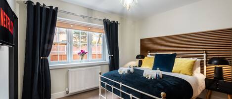 Un dormitorio con estructura de cama metálica blanca, colcha azul oscuro y ventana con cortinas negras.