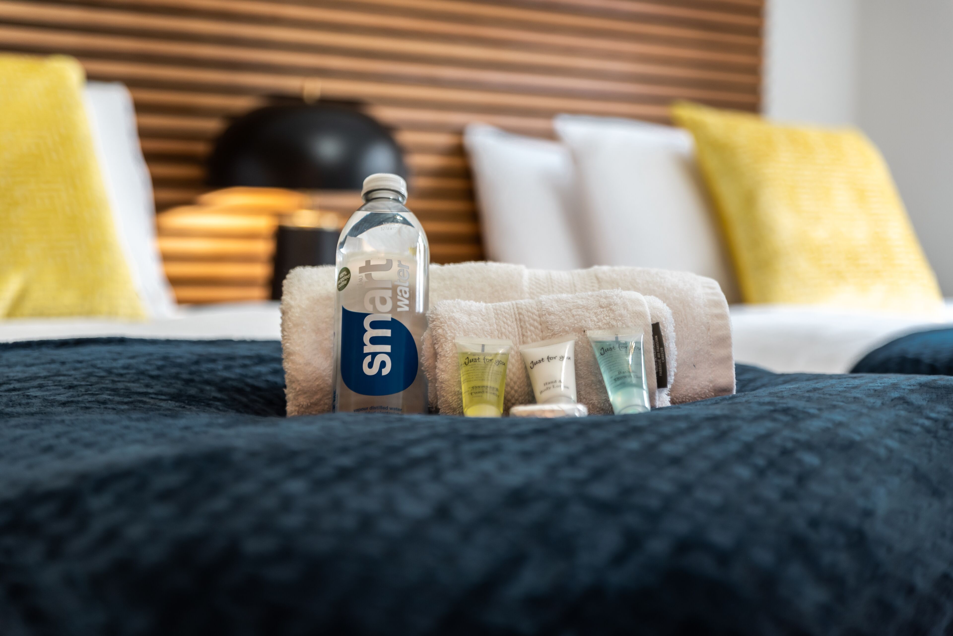Ordentlich angeordnete Pflegeprodukte auf einem Bett mit dunkelblauer Decke, einem Holzkopfende und gelben Kissen.